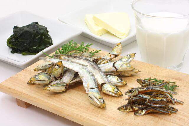 カルシウムが多く含まれる牛乳・乳製品、小魚、海藻の写真。カルシウムは、骨や歯の主要な構成成分になるほか、細胞の分裂・分化、筋肉収縮、神経興奮の抑制、血液凝固作用の促進などの働きがあります。一日に必要な食事摂取基準は、国民栄養調査の摂取量、腸管からの吸収率、骨代謝)、尿中排泄を考慮し定められています。