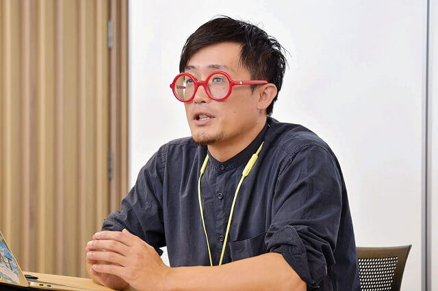 写真1、日高村役場企画課主幹の安岡周総さんの写真。「スマホで困ったら『赤い眼鏡の職員』といつでも役場に電話を」と抜群の親しみやすさで応対している。