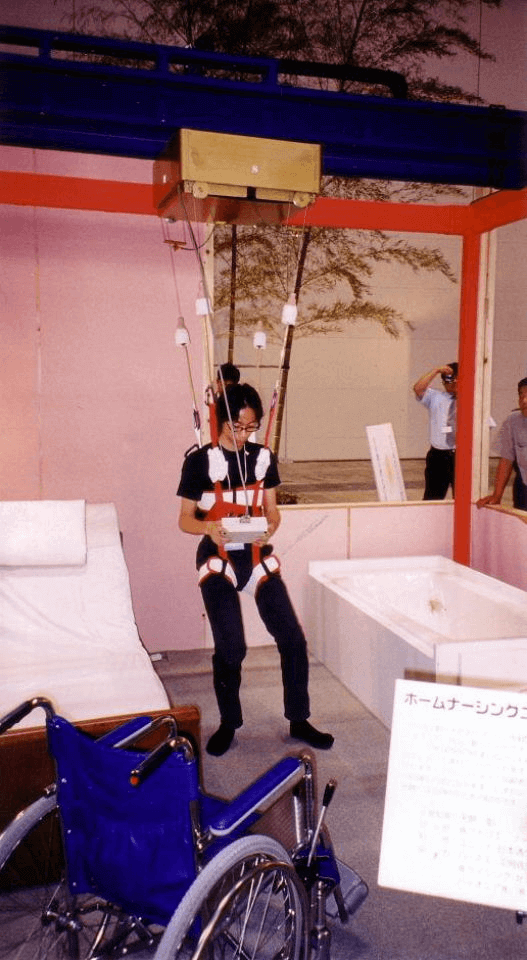 写真:男性が天井に設置されたクレーンから伸びたハーネスを身体に装着し、リモコンを操作しながらベッドから浴槽まで移動している写真