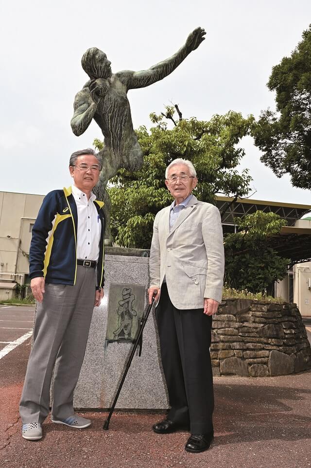 写真2：大阪市長居障がい者スポーツセンターの正門前にある片足で砲丸投げをするブロンズ像の前で撮影された小山館長と藤原さんのツーショット写真。