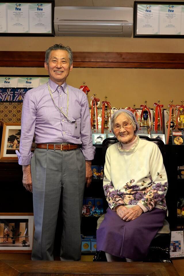写真5：長岡さんと長男の宏行さんのツーショット写真。背後には、獲得したメダルと能の舞台写真が飾られている様子を表す。