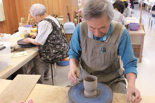 写真3：いきがいセンター内で販売するいきがい焼きを作陶する高齢者の写真。手回しのろくろで陶芸に集中する様子を表す。