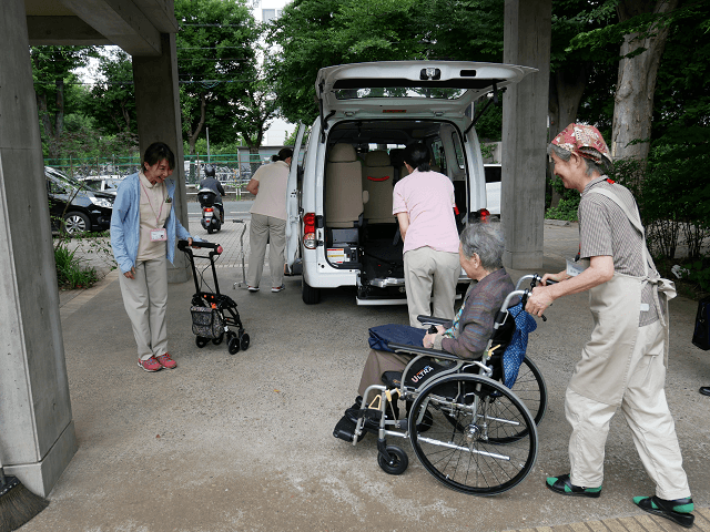 高齢の女性が乗った車椅子をボランティアの女性が押して施設に向かう写真