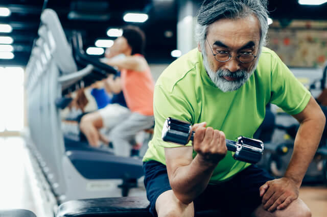 トレーニングジムでダンベルアームカールを行っている高齢男性の写真。行動体力の中でも健康関連体力（全身持久力、筋力、柔軟性、ADL、身体組成）が健康を保つ上で極めて大切な体力であるとの認識から特に高齢者では筋持久力の向上を目指し、筋力トレーニングが推奨されていることを示す。