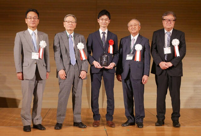 豆野智昭氏の表彰式の様子を表す写真。